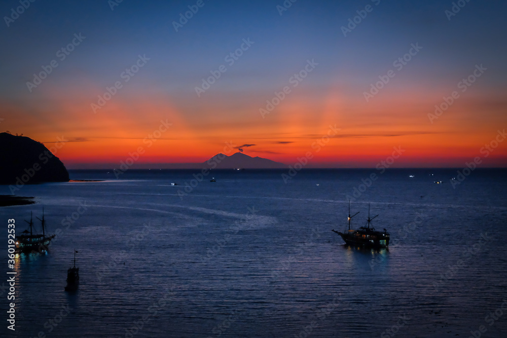A sailing boat towards a silhoutte of a mountain in the background in Labuan Bajo after sunset. Blue sky and slightly orange rays from sun is seen in the horizon