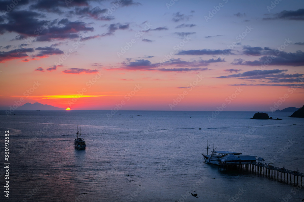 A sailing boat towards the sun, during a sunset with a mountain in the background with a marina in Labuan Bajo A sailing boat towards the sun, during a sunset with a mountain in the background with a marina in Labuan Bajo