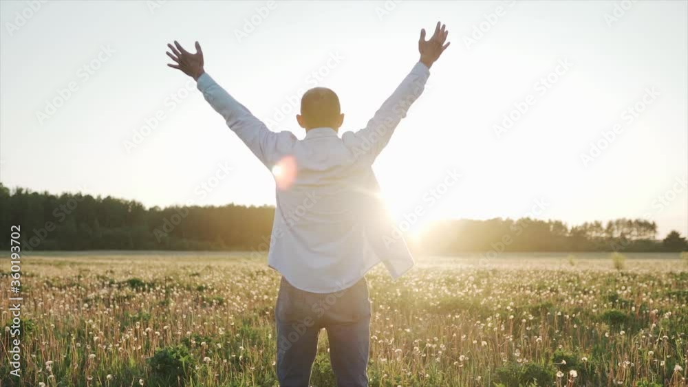 A caucasian man standing on the beautiful field during summer sunset, his hands are raised. A man look at the sky and pray. A man stand with your back to the camera. Close up. A caucasian man standing on the beautiful field during summer sunset, his hands are raised. A man look at the sky and pray. A man stand with your back to the camera. Close up.