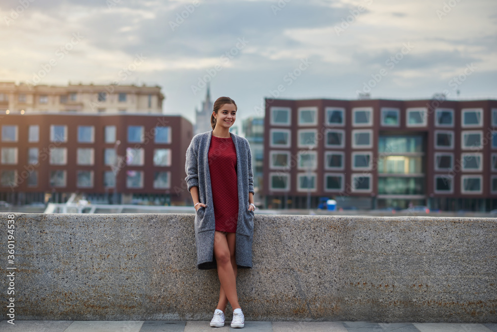 Young woman with beautiful smile dressed in cool fashionable clothes is standing near river pier against blurred old buildings with copy space during her long awaiting spring weekend to St. Petersburg