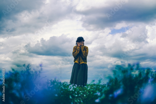 Photo A girl in a loose boho dress stands with her face in her hands in a field of flax flowers against a sky with clouds