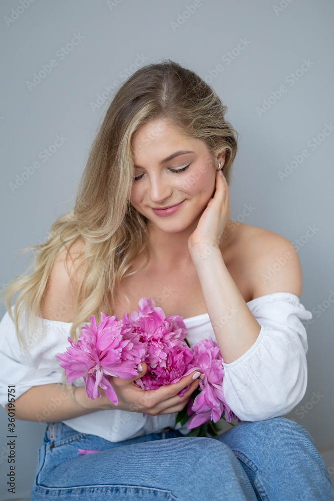 beautiful sensual girl with long blonde hair in blue jeans and a white shirt with a bouquet of pink peonies flowers. Close-up portrait of a woman in studio on a gray background.concept of feminity