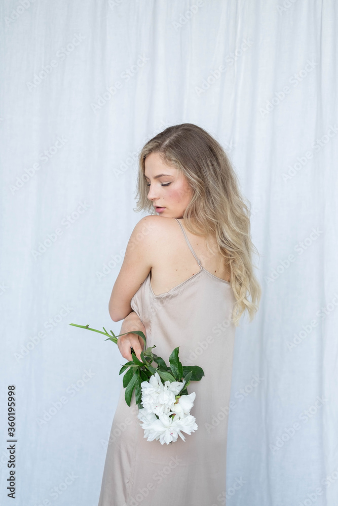 beautiful sensual girl with long blonde hair in dress with a bouquet of pink peonies flowers. Close-up portrait of a woman in studio on a gray white background.concept of feminity spa