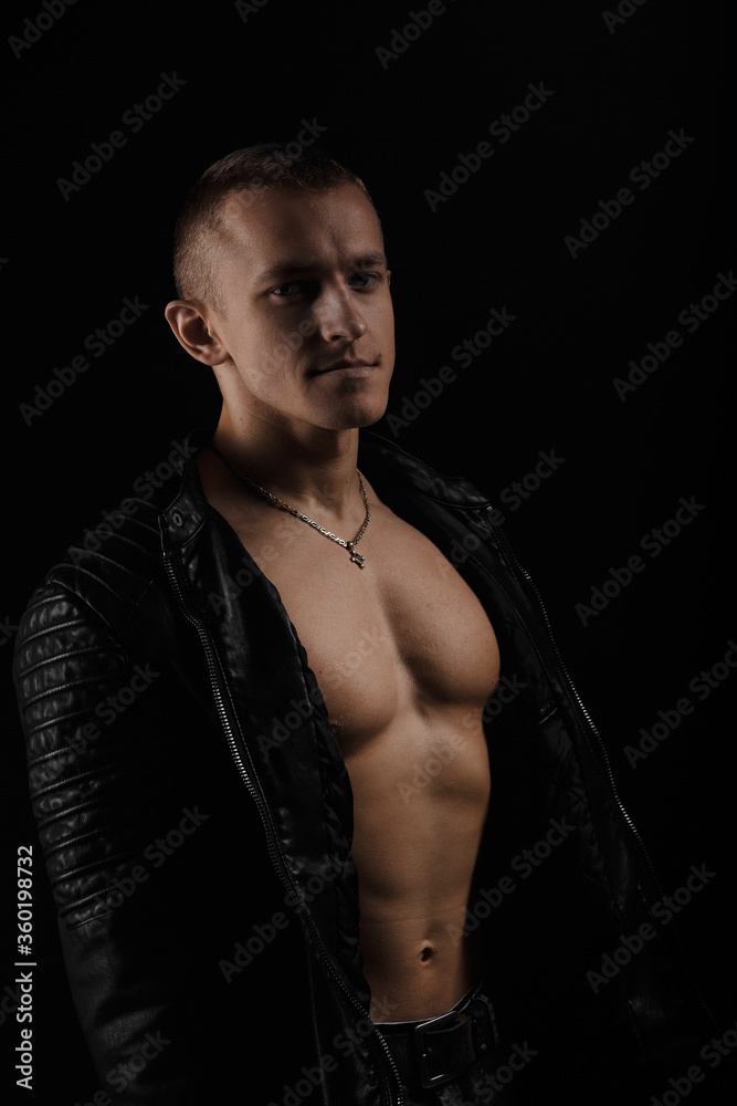 Fototapeta premium Handsome man wearing leather jacket on a naked muscular torso, on a dark background, looking into the camera.