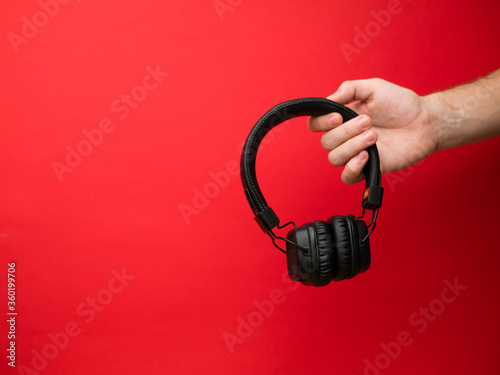 A young woman holds a pair of black wireless headphones in her hand. Hand of a girl with headphones on a red background. Musical concept.