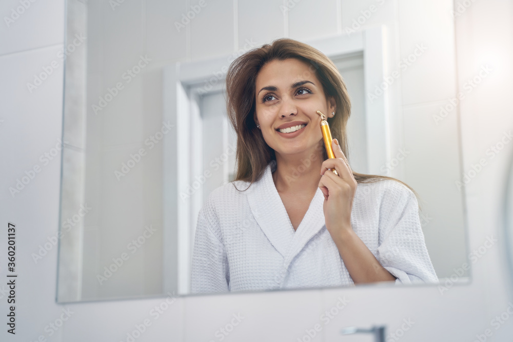 Joyful young woman using golden facial massager in bathroom Joyful young woman using golden facial massager in bathroom