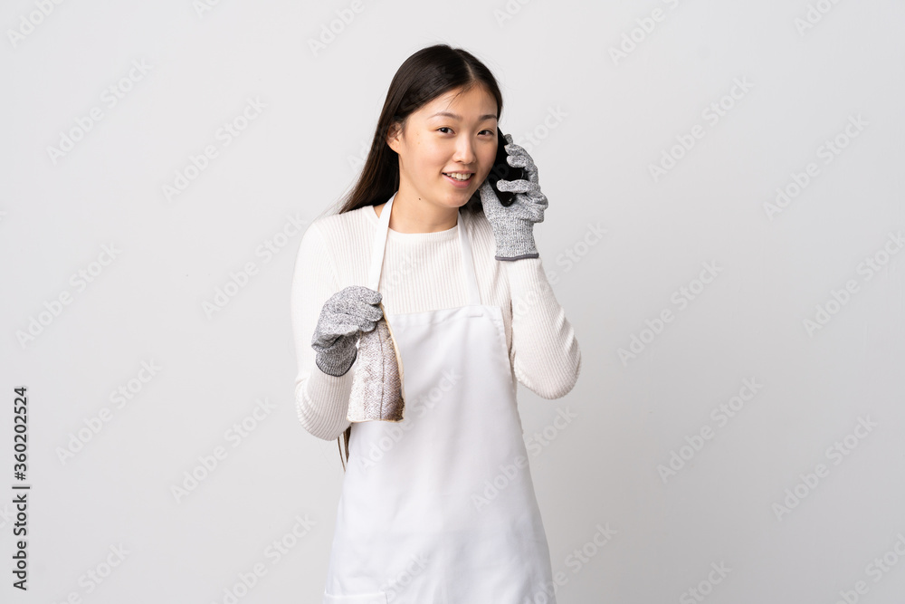 Chinese fishmonger wearing an apron and holding a raw fish over isolated white background keeping a conversation with the mobile phone with someone Chinese fishmonger wearing an apron and holding a raw fish over isolated white background keeping a conversation with the mobile phone with someone