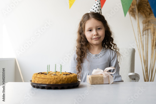 A caucasian girl has a holiday in the house. She is smart with her hair loose and in a festive hat. A child sits at a table on which stands a cake with candles 