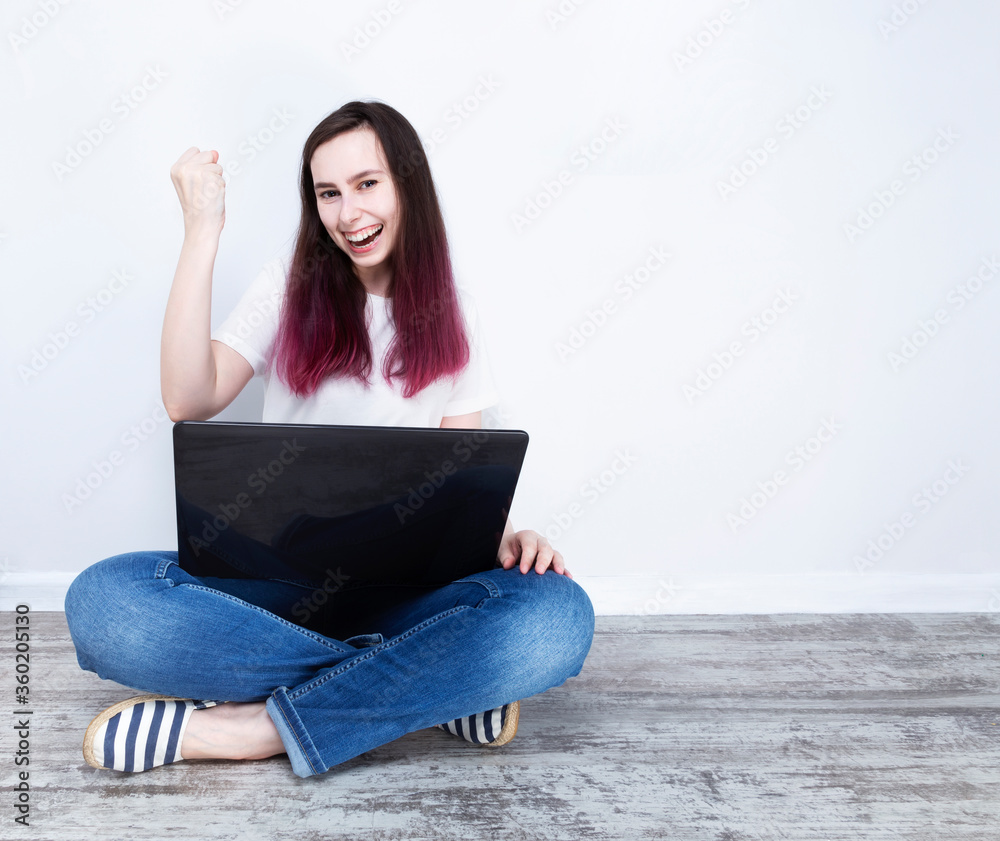 Naklejka premium Young woman sitting on floor with laptop victoriously raises hands, grey background. Copy space