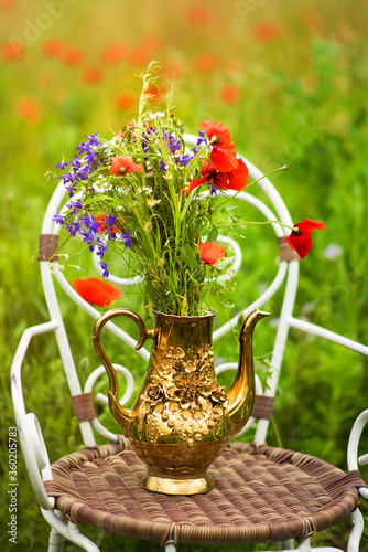Vertical picture of bouquet from fresh wild flowers, chamomile and poppies, in golden teapot on chair in middle of meadow with poppies and green grass. Rural still life