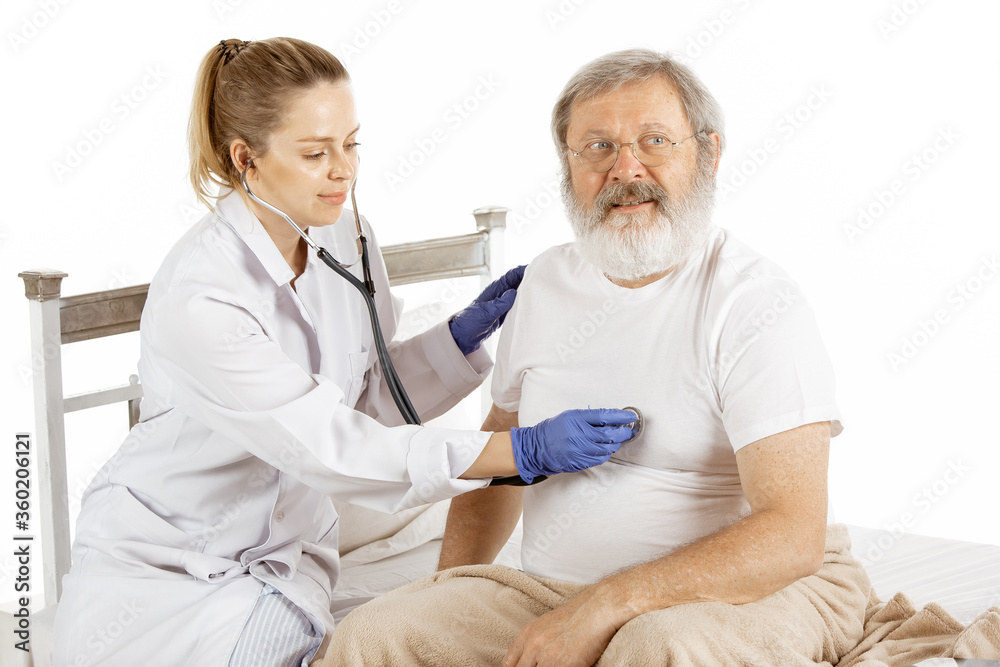 Obraz premium Elderly old man recovering in a hospital bed isolated on white background. Getting care. Concept of healthcare and medicine. Nurse listen with stethoscop for treatment and fast diagnosting. Copyspace.