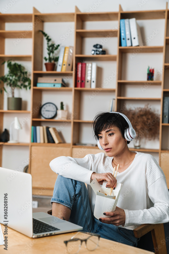 Image of serious woman eating asian noodles while working with laptop Image of serious woman eating asian noodles while working with laptop