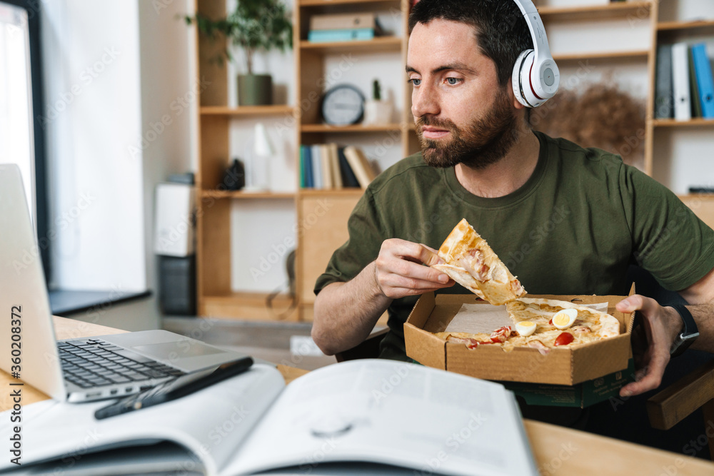 Photo of focused bearded man eating pizza while working with laptop Photo of focused bearded man eating pizza while working with laptop