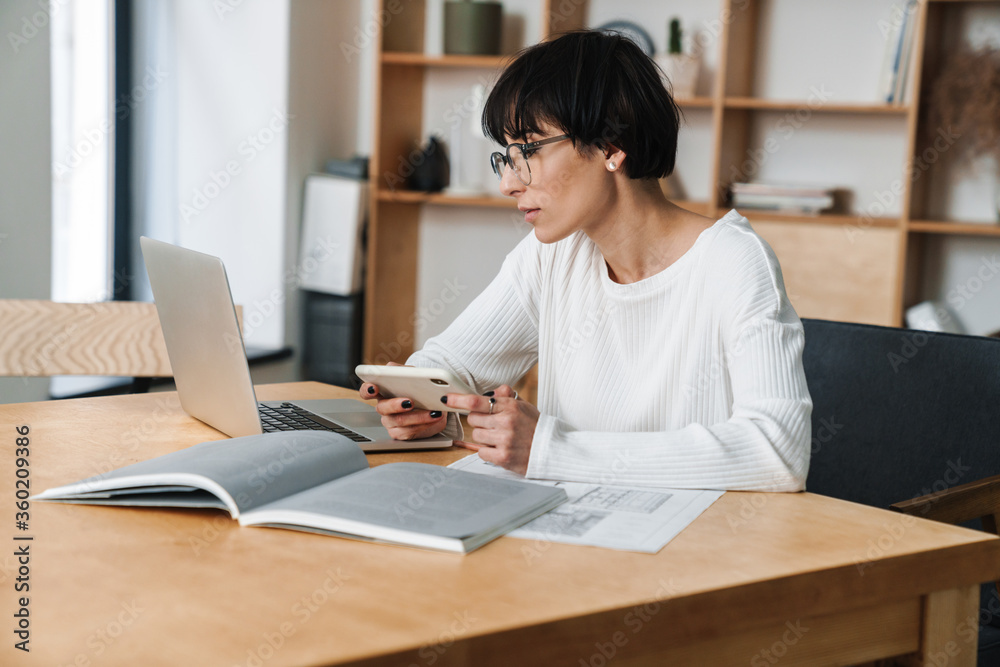 Image of focused woman in eyeglasses working with laptop and cellphone Image of focused woman in eyeglasses working with laptop and cellphone