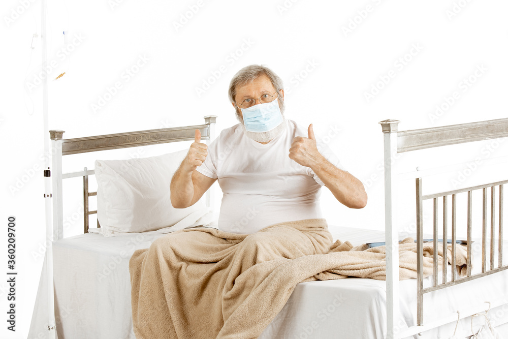 Elderly old man recovering in a hospital bed isolated on white background. Getting care and treatment. Concept of healthcare and medicine, diagnostics. Put on protective face mask. Copyspace.