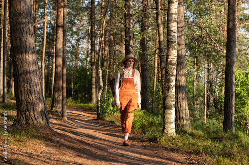 Woman botanist in orange overalls with backpack on ecological hiking trail in forest. Naturalist exploring wildlife and ecotourism adventure walking in a wildlife national park