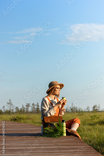 Woman botanist with backpack sitting on wooden path through peat bog swamp in wildlife national park. Naturalist resting on boardwalk, drinking tea, enjoying the moment at sunset. Ecotourism 