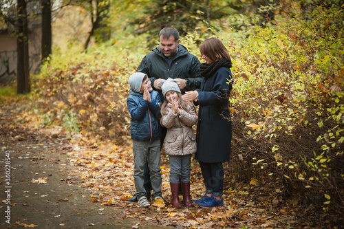 A cute family is standing in the autumn park with orange foliage. The wife lies on her husbands chest, Daughter hugs her mother, the son is standing next to him. Beautiful portrait, soft focus.