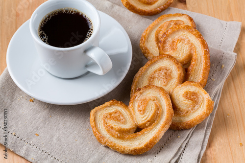 A cup of dark hot black coffee beside pile of palmier biscuits dropped around on a wood table