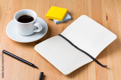 A light wood desktop table with a cup of coffee  a pen  a open notebook  and some colour note paper around
