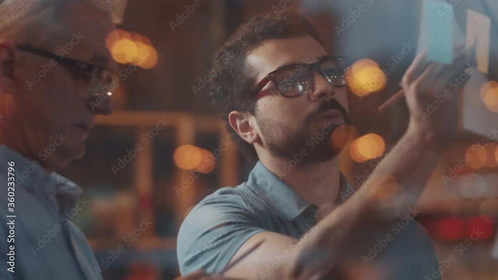 Young middle eastern businessman and his elderly Caucasian colleague speaking and writing plans on adhesive notes attached to glass wall in office; bokeh lights reflected in glass