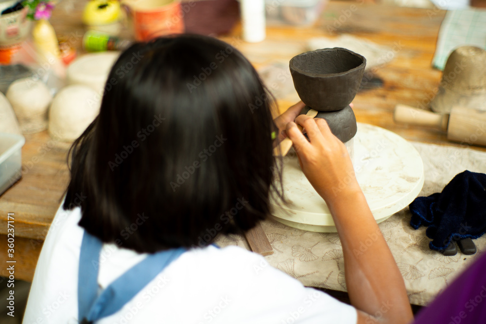 Closeup hands of little girl molding the clay on the wheel tray by wooden stick with blurred back portrait of girl in foreground