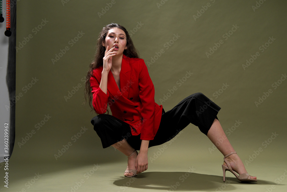A sensual young woman in a red jacket, culottes and shoes with wet long black hair poses against a green background in the studio. A sensual young woman in a red jacket, culottes and shoes with wet long black hair poses against a green background in the studio.
