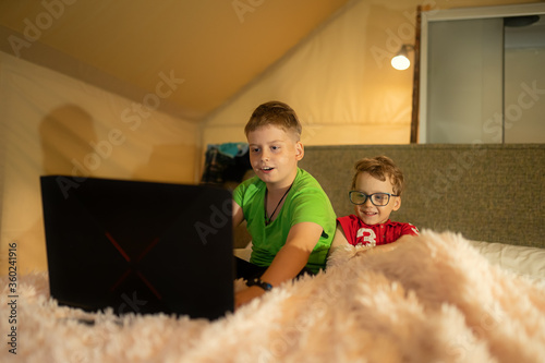 Two children watch cartoons on a laptop while lying on a bed in a tent house