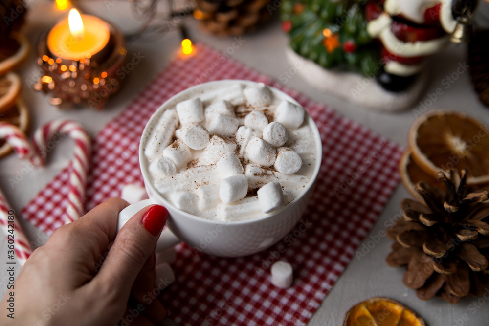 
Female hand holds a cup with hot chocolate and marshmallows on a table decorated for Christmas