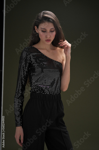 A beautiful young tall girl with long black hair poses in a shiny blouse and black culottes on a green background. Buty and fashion photo shoot.