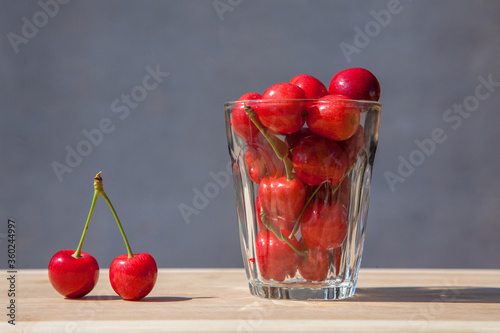 Two ripe red cherry berries and a glass cup full of berries on a gray background