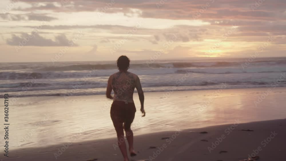 Handheld tracking of young man with tattoo on his back running towards ocean and doing somersault on coast on Bali at sunset Handheld tracking of young man with tattoo on his back running towards ocean and doing somersault on coast on Bali at sunset