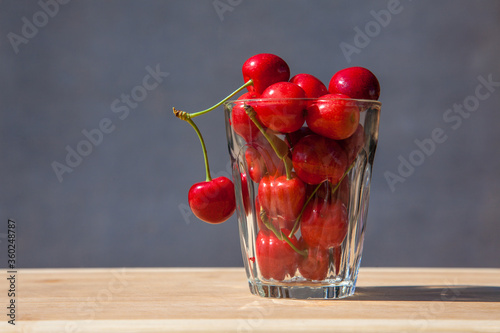 Two ripe red cherry berries hanging on a glass glass full of berries on a gray background