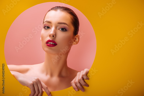 naked beautiful woman with crimson lips in yellow paper circle hole on pink background