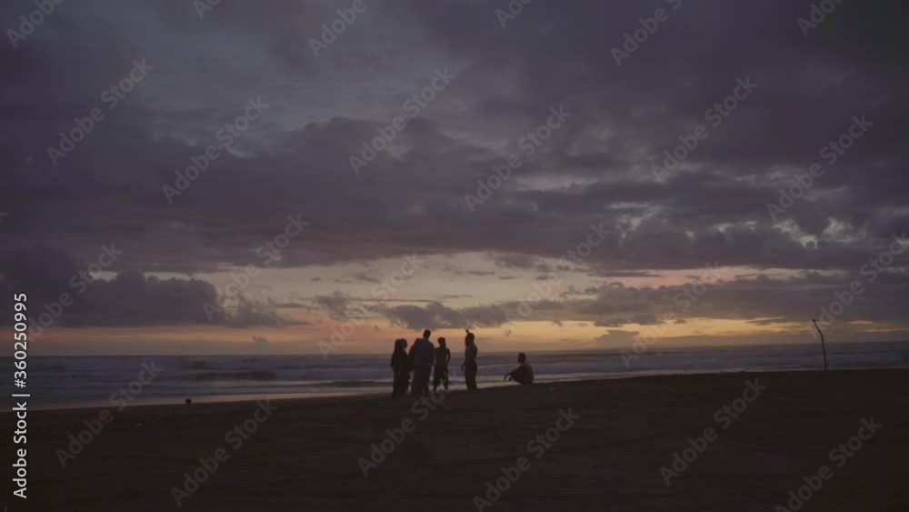 Wide tracking shot of silhouettes of young people hanging out together on beach on coast of Bali at dusk Wide tracking shot of silhouettes of young people hanging out together on beach on coast of Bali at dusk