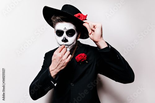 White male model in black hat posing with skull makeup. Indoor shot of joyful zombie guy having fun in day of the dead.