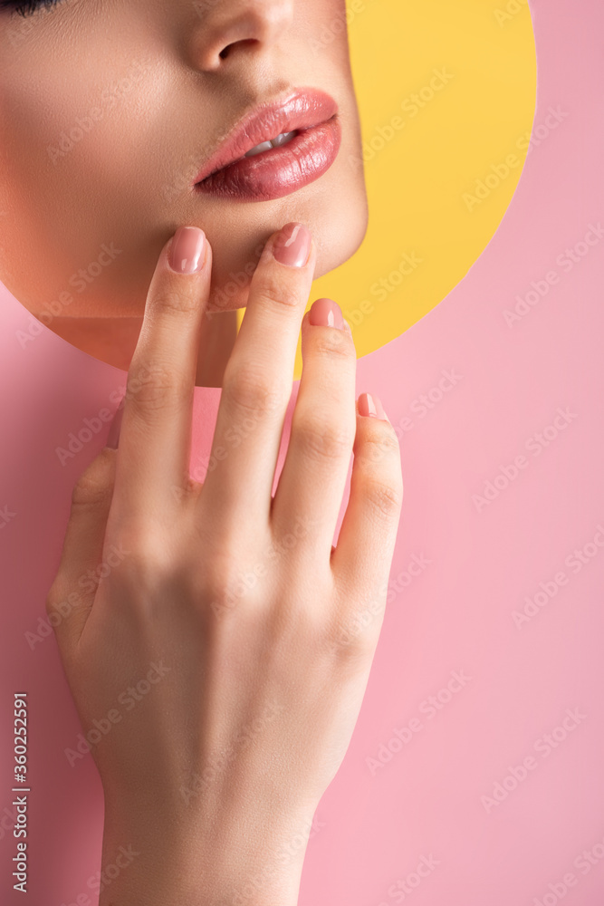 partial view of beautiful woman with shiny lips in pink paper round hole with hand isolated on yellow