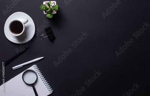 White cup with coffee, pens, notebook with pen and flower in a pot on a black background. View from above. A place to copy. Business concept