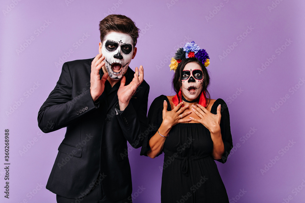 Shocked caucasian woman with black hair posing in halloween costume. Surprised zombie guy standing on purple background with girlfriend. Shocked caucasian woman with black hair posing in halloween costume. Surprised zombie guy standing on purple background with girlfriend.