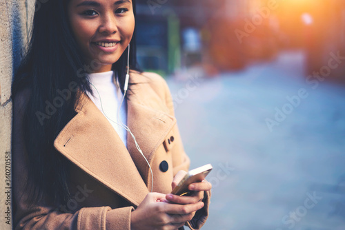 Charming cheerful attractive young female hipster girl standing outdoors listening to favorite songs via earphones while holding modern cellular standing on copy space area for advertising content