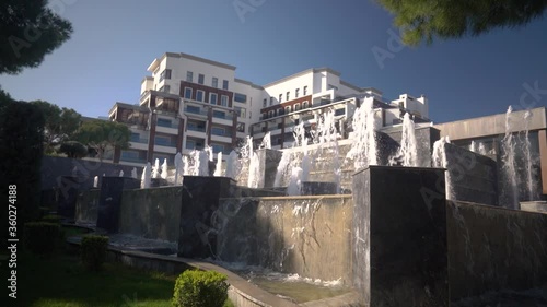 A white fountain with water splashes. Water flows over large artificial stones. Modern building in background. 