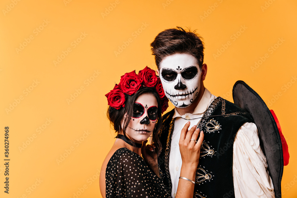 Obraz premium Graceful mexican woman standing near man with black sombrero. Indoor portrait of couple in masquerade costumes.
