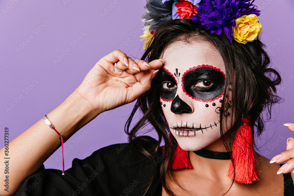 Close-up portrait of refined woman with dark eyes posing in masquerade costume. Cute latin lady in flower wreath preparing for halloween.