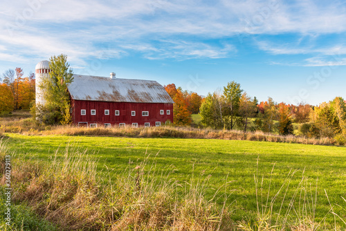 Idyllic rural landscape with an old red wooden barn with a silo under blue sky at sunset. A meadow is in foreground and colourful autumn trees are in background. Countryside of Vermont, United States.