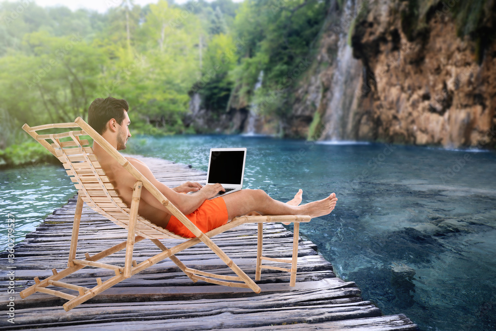 Young man with laptop relaxing on sun lounger near lake and waterfall. Luxury vacation