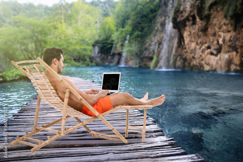 Young man with laptop relaxing on sun lounger near lake and waterfall. Luxury vacation