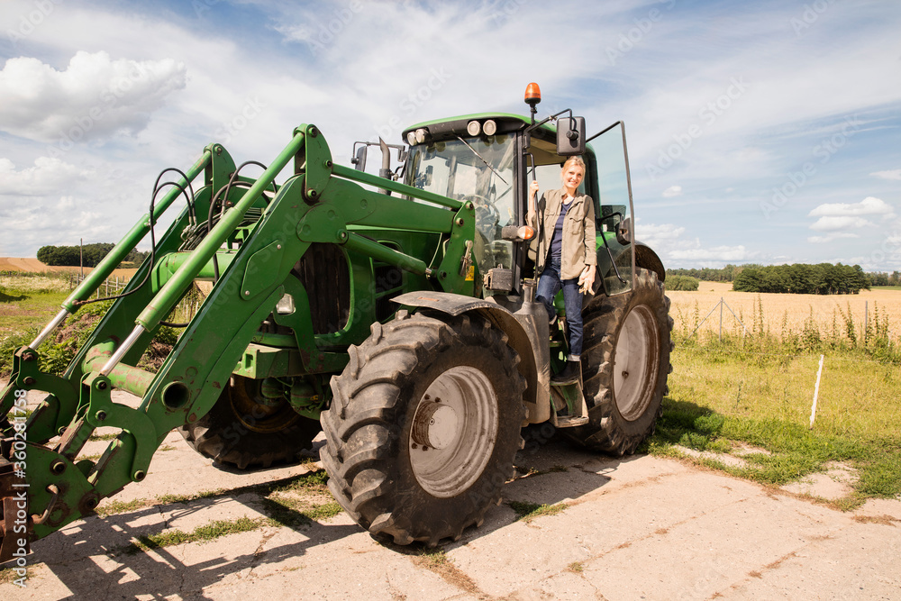 Full length of smiling mature woman standing on agricultural machinery against sky Full length of smiling mature woman standing on agricultural machinery against sky