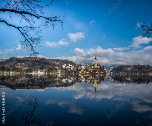 Reflection of lake Bled island church tower and hills covered with trees in calm water. Famous tourist landmark in Slovenia. Winter season under Alps mountains. Low angle  wide shot