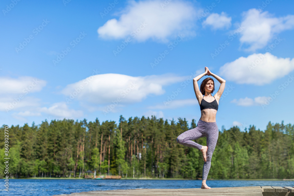 Woman practicing yoga on a lake standing on log in yoga tree pose. fitness lifestyle at the outdoors nature background. Sunny day. copy space 