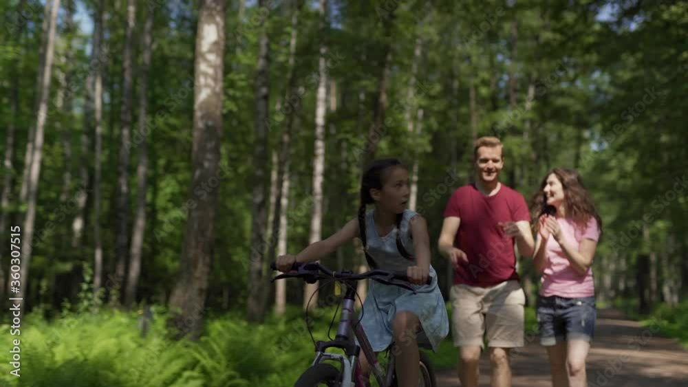 Caring parents teaching little daughter to cycle down trail in green park. Excited girl enjoying learning how to ride on bike Caring parents teaching little daughter to cycle down trail in green park. Excited girl enjoying learning how to ride on bike